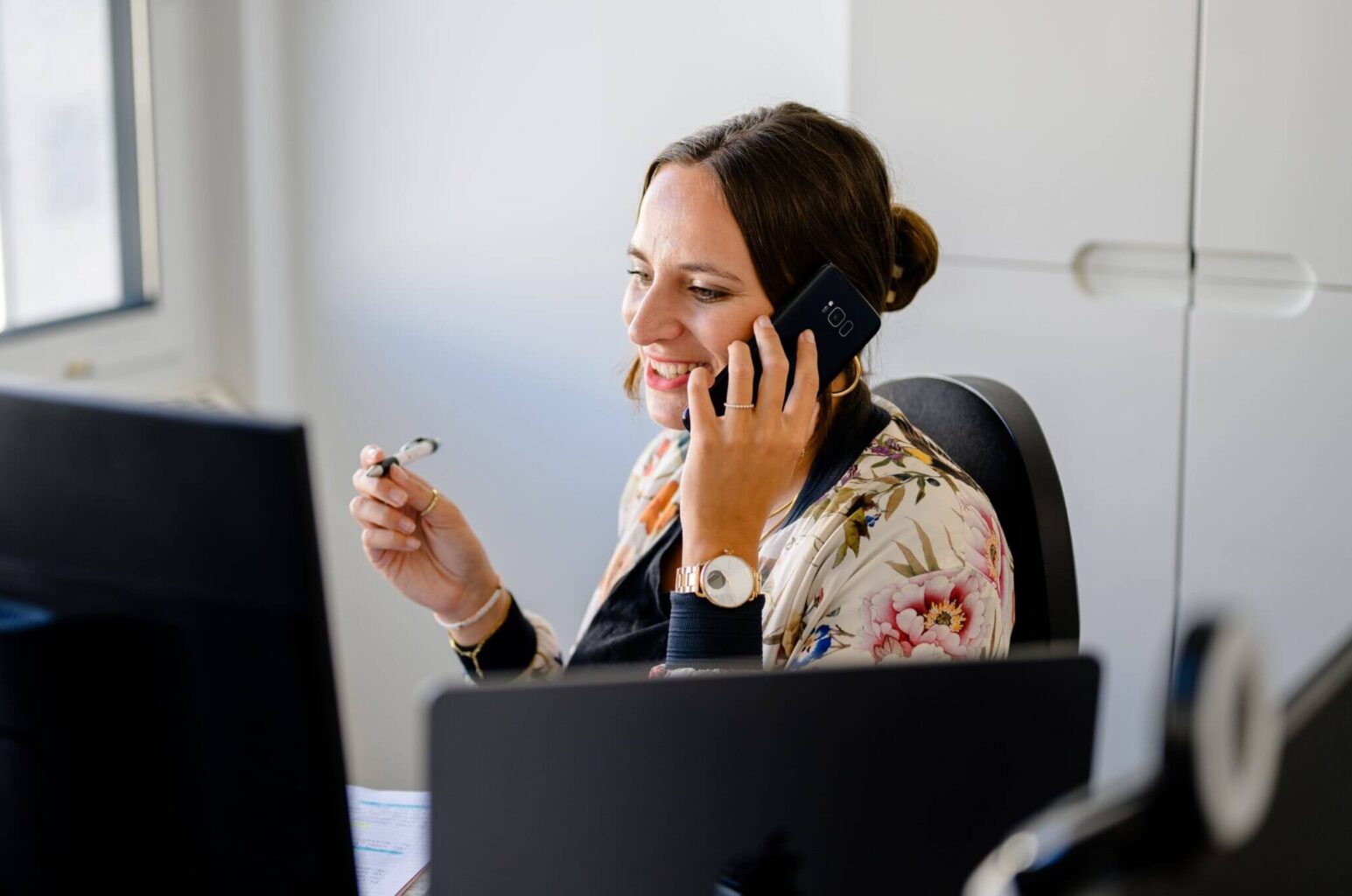 Lächelnde Frau am Schreibtisch bespricht SEO-Konzept auf dem Smartphone, hält Brille, mit Computer und Tageslicht aus dem Fenster.