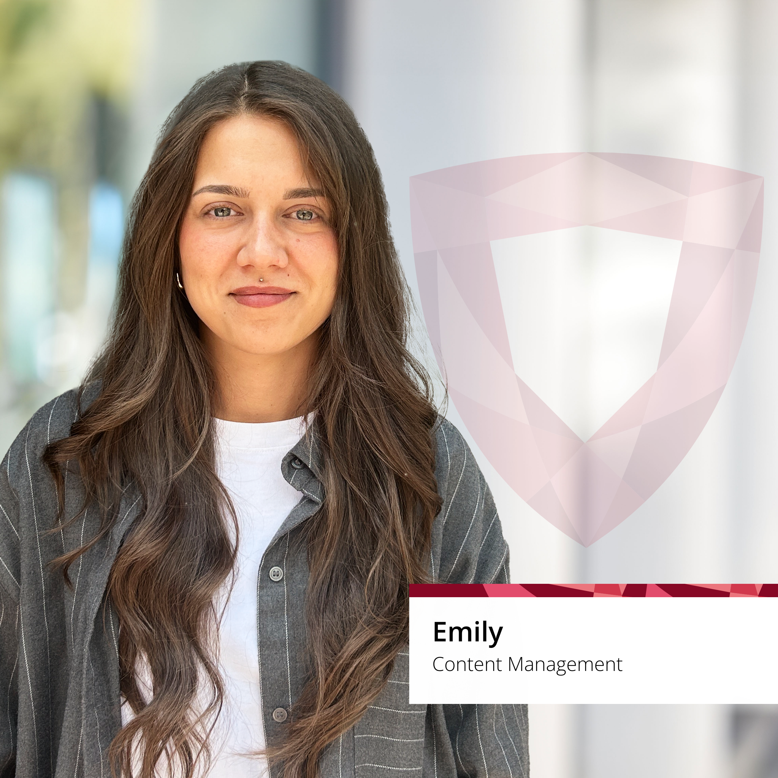 Frau mit langen braunen Haaren in gestreiftem Hemd lächelt im Freien; transparentes Schild und Namensschild „Emily, Content Management“ neben ihr.